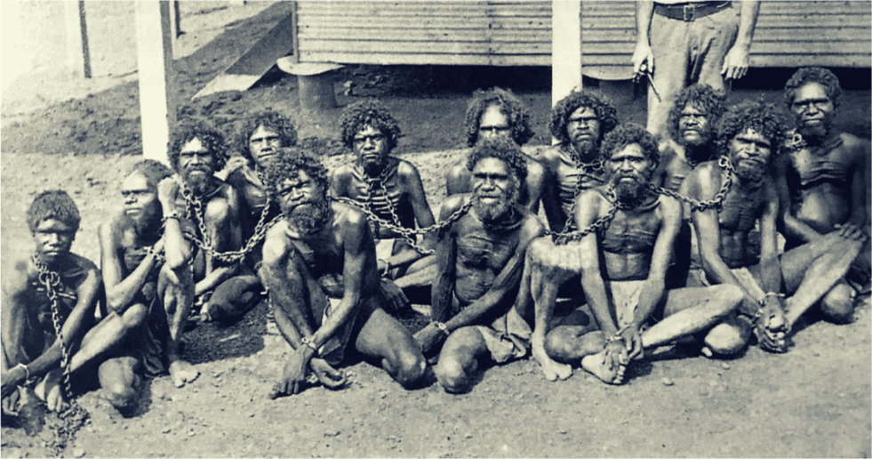 A black and white image of a group of Aboriginal people sitting with chains around their necks