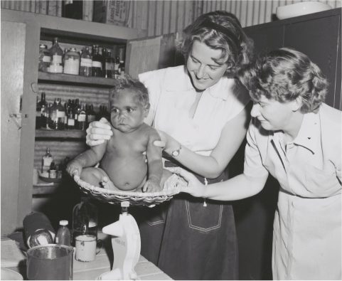 A black and white photo of Sisters Nemertothy and Page weighing an Aboriginal baby, Hooker Creek Mission, Northern Territory, 1958